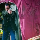 Ein Mann zielt mit einer Pistole unter einem roten Vordach. Im Hintergrund sieht man Obstkisten und eine Person in Blau.