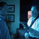 Ich kann den Text nicht erkennen. Zwei Männer in einem Raum, einer hält eine Pistole, die Beleuchtung ist blau.