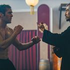 Zwei Männer im Sparring, der eine muskulös und oberkörperfrei, der andere in dunkler Kleidung, asiatische Kampfsporthalle.