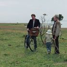 Ein Mann mit Fahrrad, ein älterer Mann und ein Kind mit Hut stehen auf grünem Feld nahe einem See.
