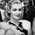 Ich kann keine Texte oder Personen beschreiben. Eine blonde Frau lächelt, trägt Blumen im Haar und ist elegant gekleidet.
