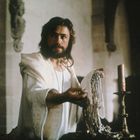 Ein Mann mit Bart in einem weißen Gewand hält Schmuck in einer historischen Umgebung mit Kerzenständern.
