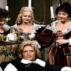 Drei Frauen in barocken Kleidern umgeben einen Mann auf einem Sofa in einem historischen Raum mit Kerzenständern.