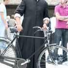 Ein Mann in einem schwarzen Priestergewand und Baskenmütze steht neben einem Fahrrad.