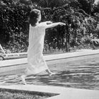 Eine Frau im weißen Kleid steht scheinbar auf einem Pool. Im Hintergrund sind Pflanzen und eine Sonnenliege zu sehen.