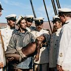 Sieben Männer in weißen Marineuniformen stehen auf einem Schiff, einer hält einen bewusstlosen Jungen.