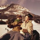 Zwei Personen in Winterkleidung sitzen lächelnd in einer verschneiten Landschaft mit Bergen im Hintergrund.