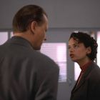 Ein Mann und eine Frau in grauen Anzügen stehen sich gegenüber in einem Büro, sie schaut ernst, er hat den Rücken zur Kamera.