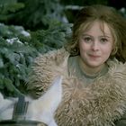 Ein junges Mädchen lächelt, umgeben von schneebedeckten Tannen, mit einem weißen Pferd im Vordergrund.