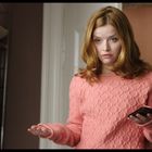 Eine Frau mit roten Haaren trägt einen rosa Pullover und hält ein Smartphone in der Hand, stehend in einem Zimmer.