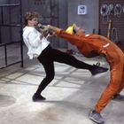 Eine Frau in Weiß stößt einen Mann in orangefarbener Arbeitskleidung zurück. Beide scheinen in Bewegung zu sein.