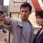 Zwei Personen stehen nebeneinander. Der Mann trägt eine Waffe, die Frau schaut ernst. Im Hintergrund sind Gebäude zu sehen.