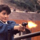 Eine Frau in blauer Jacke schießt mit einer Pistole, während ein Feuerstrahl aus der Waffe zu sehen ist.