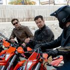 Drei Männer auf orangen Motorrädern. Zwei tragen Lederjacken, einer einen Helm. Sie stehen vor einem Betonhintergrund.