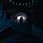 Keine erkennbaren Texte. Dunkle Treppe in einem großen Raum, eine Gestalt steht oben vor einem runden Fenster.