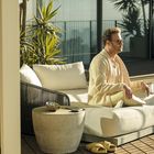 Ein Mann sitzt entspannt auf einem Sofa auf einer Terrasse. Er trägt helle Kleidung und eine Sonnenbrille, umgeben von Pflanzen.