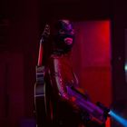 Ein Mensch in einem roten Anzug mit Gitarren auf dem Rücken und einem großen Gewehr in der Hand, in einer düsteren Umgebung.
