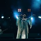 Eine Sängerin in einem fließenden Kleid performt auf einer Bühne mit zwei Gitarristen und bunten Lichtern.