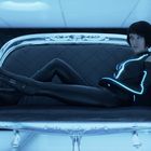 Eine Person in einem schwarzen Anzug mit leuchtenden blauen Linien sitzt auf einem transparenten Sofa in futuristischer Umgebung.