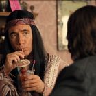 Zwei Personen sitzen in einer Bar; eine trägt ein indianisch inspiriertes Outfit und trinkt einen Cocktail mit Erdbeere.