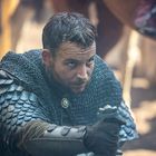 Ein Mann in Kettenhemd und Schuppenpanzer hält ein Schwertgriff. Sein Gesicht wirkt konzentriert und entschlossen.