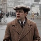 Ein Mann blickt besorgt in die Ferne. Er trägt einen braunen Mantel und eine Schiebermütze. Im Hintergrund verschwommene Stadt.