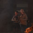 Zwei Frauen stehen zusammen, eine hält einen kleinen Hund. Sie sehen besorgt aus, im Vordergrund sind Flammen sichtbar.