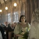 Eine Frau in festlichem Kleid hält Blumen, umgeben von Menschen in einer historischen Kirche mit hohen Decken und steinernen Bögen.