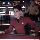 Person in roter Uniform mit Abzeichen, schaut ernst. Hintergrund: futuristisches Kontrollzentrum mit Bildschirmen und Menschen.