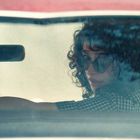 Keine Texte. Eine Frau mit lockigem Haar und Sonnenbrille schaut über die Schulter aus einem Auto.