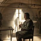 Ein Mann in Militäruniform sitzt in einem düsteren Raum, durch ein Fenster fällt Licht. Personen im Hintergrund.