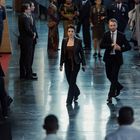 Zwei Personen in Business-Kleidung gehen durch eine große, elegante Halle mit verschiedenen Menschen im Hintergrund.
