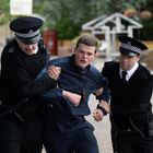 Zwei Polizisten führen einen Mann, beide tragen Uniformen und Mützen, während sie den Mann an den Armen festhalten.