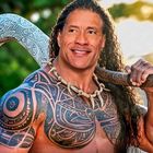 Eine Person mit langem Haar und tribalem Tattoo trägt einen großen Hacken. Hintergrund: verschwommenes Grün.