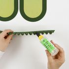Text auf der Klebeflasche: "BASTELKLEBER MULTI GLUE". Hände kleben grüne Pappstreifen mit Zickzackkanten.