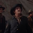 Drei Männer in Westernkleidung schauen aufmerksam, ein Mann hält Pferde vor einer felsigen Landschaft.