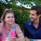 Eine Frau mit pinkem Haar und Tattoos sitzt neben einem Mann. Sie haben entspanntes Gespräch im Freien unter Bäumen.