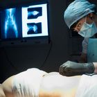 Ein Chirurg mit Mundschutz und Haube führt eine Operation durch, Röntgenbilder sind im Hintergrund zu sehen.