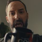 Unklarer Text auf schwarzer Tasse. Ein Mann mit Bart schaut überrascht oder verärgert. Links sind Jalousien sichtbar.