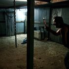 Kein Text vorhanden. Eine dunkle Scheune mit Wellblechwänden. Ein Mann hält ein Messer, eine Frau steht verängstigt.