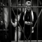 Ein Priester mit Kreuz, hinter Gittern stehend, zusammen mit einer umarmenden Person und einem Soldaten links.