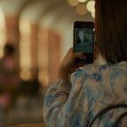 Eine Person macht ein Foto mit einem Smartphone in einem Innenraum. Sie trägt ein blau gemustertes Oberteil.