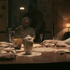 Eine Gruppe von fünf Menschen sitzt still um einen Esstisch. In der Mitte steht eine Teekanne. Eine Person trägt eine Sauerstoffmaske.