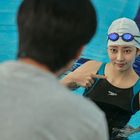 Ein Schwimmer im Pool trägt eine weiße Badekappe und Schwimmbrille, zeigt auf sich selbst und trägt einen Anzug mit "speedo".