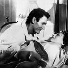 Ein Mann beugt sich lächelnd über eine Frau in einem Bett. Sie schauen sich liebevoll an. Es ist eine schwarz-weiße Szene.