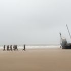 Ein Strand bei Nebel. Links marschierende Soldaten, rechts ein gestrandetes blaues Schiff. Wellen im Hintergrund.