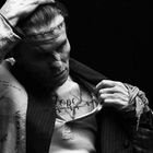 Mann zieht Hemd zurück, um ein Tattoo mit dem Wort "Hope" auf der Brust zu zeigen, mit Narben auf Stirn und Kleidung.