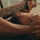 Eine Frau liegt nahe einem Gitarrenspieler. Ihr Gesicht wird sanft gehalten, harmonische Atmosphäre.