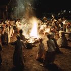 Menschen tanzen in einem Kreis um ein großes Lagerfeuer. Die Szene wirkt lebendig und festlich.