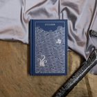 Der Text "STEELBORN" ist auf einem blauen Buchcover. Wellen und ein Schiff sind kunstvoll eingeprägt. Stoff und Schwert liegen daneben.